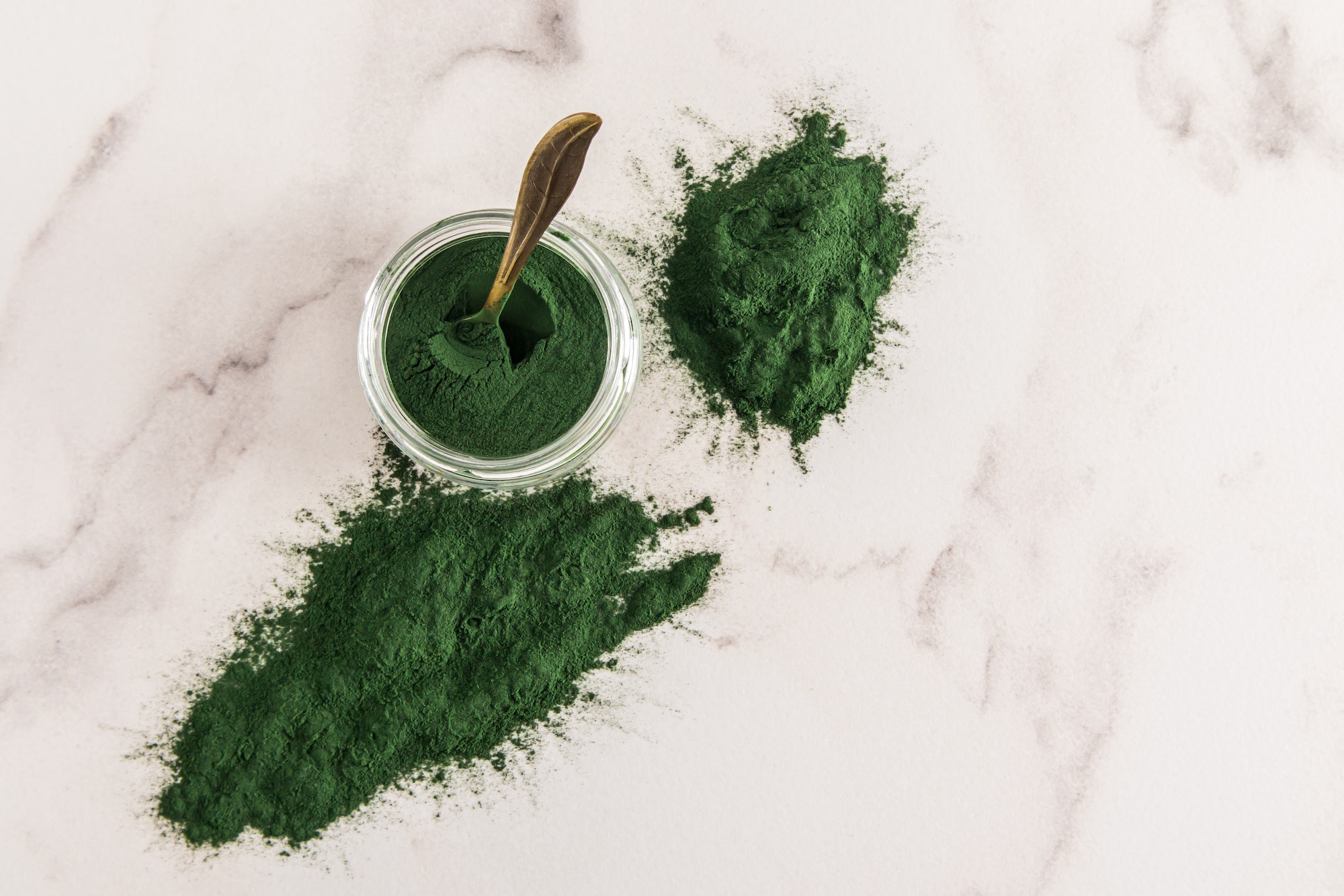 scattered spirulina powder on a marble white background and a glass jar with an organic food additiv